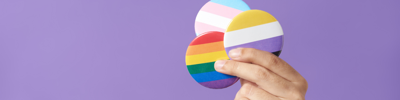 hand holding a trans flag button, a pride flag button, and a nonbinary flag button